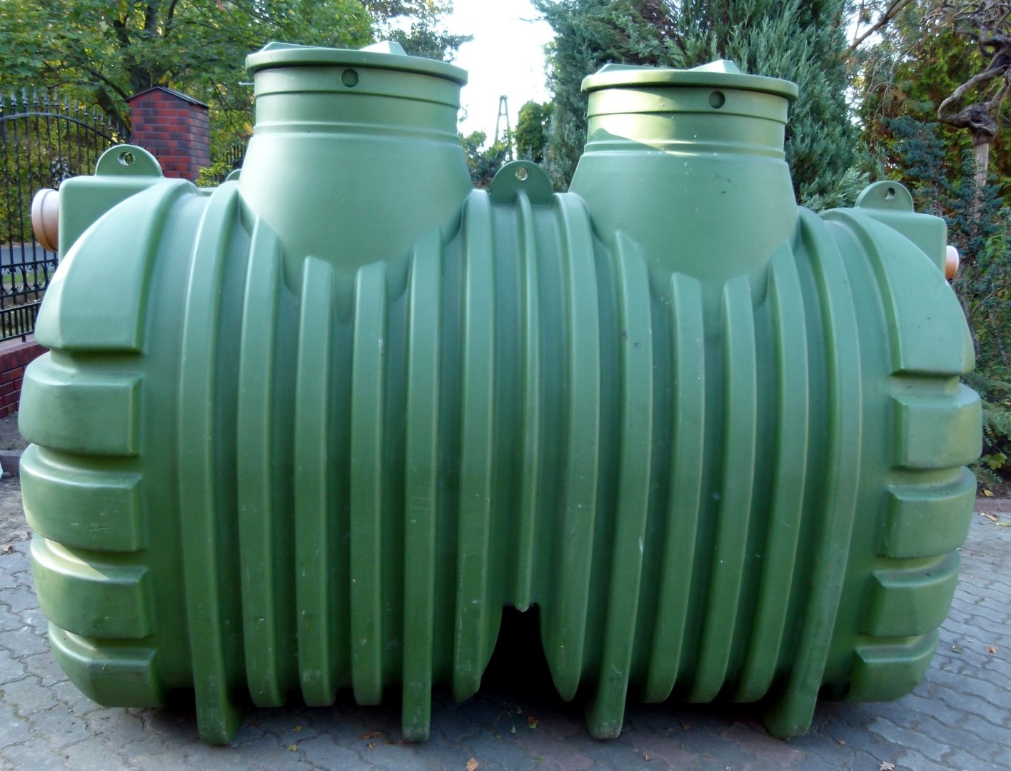 Zielony, plastikowy zbiornik dwukomorowy z wyraźnymi pionowymi żebrami, ustawiony na kostce brukowej, z widocznymi elementami montażowymi i pokrywami, na tle zieleni ogrodowej.