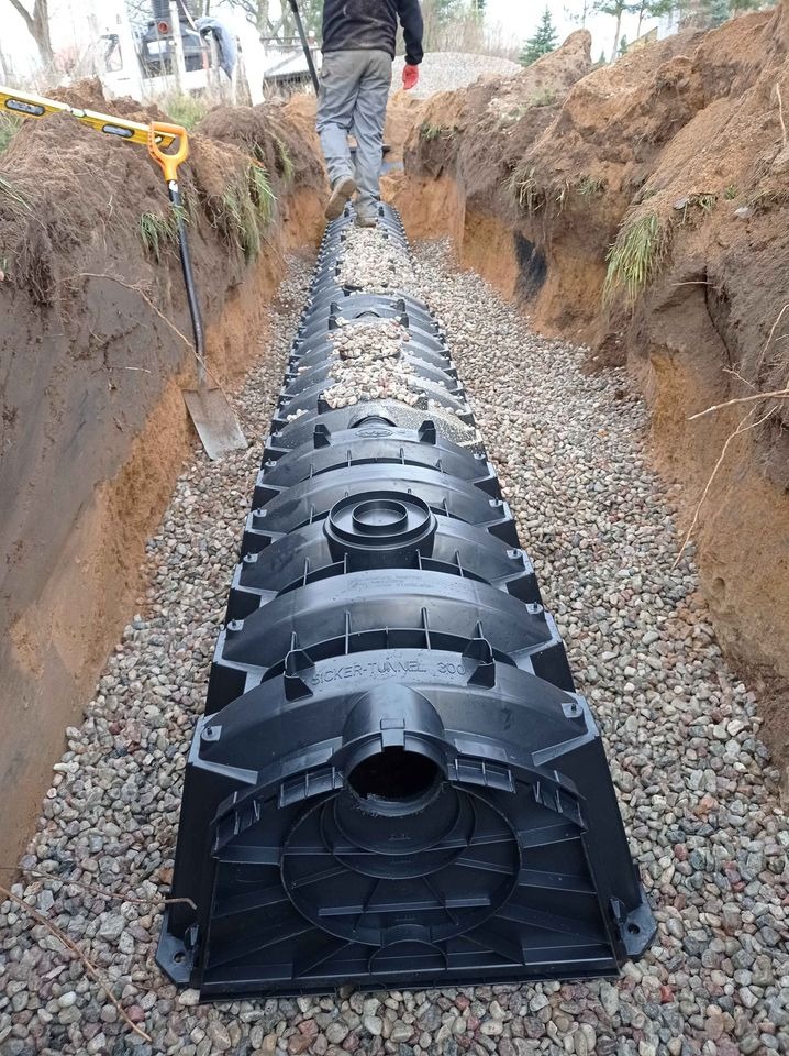 Instalacja drenażowa z tuneli rozsączających Sicker-Tunnel 300 w wykopie wypełnionym żwirem, widoczna łopata i poziomica.