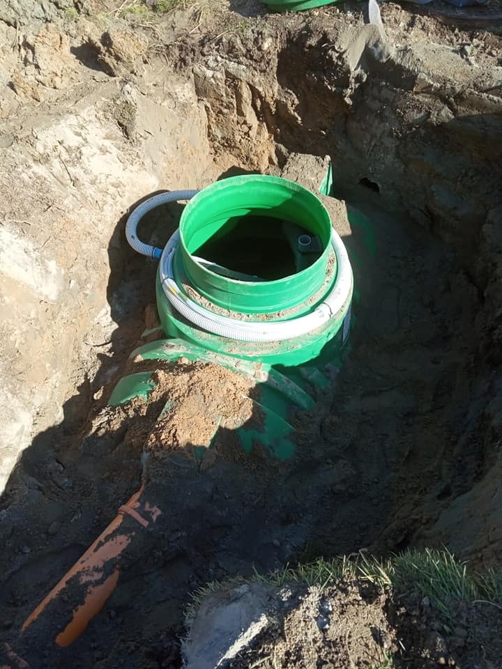 Zielony zbiornik z białymi rurami w wykopie ziemnym, widoczny fragment pomarańczowej rury kanalizacyjnej, trawa na skraju wykopu.