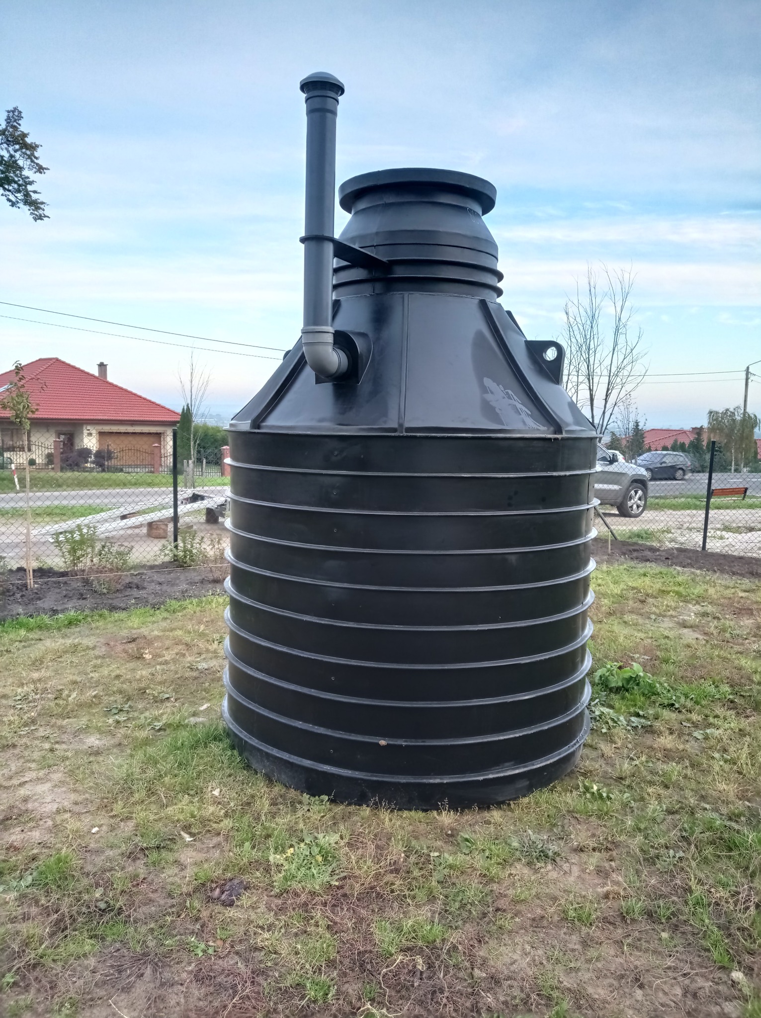 Czarny, plastikowy zbiornik na trawniku przed domem jednorodzinnym, z widoczną rurą wentylacyjną i elementami montażowymi, otoczony świeżo posadzoną roślinnością i ogrodzeniem z siatki.