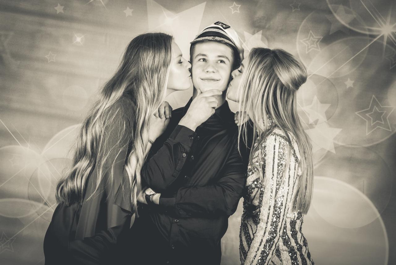 Czarno-białe zdjęcie młodego mężczyzny w czarnej koszuli i czapce kapitana, całowanego w policzki przez dwie kobiety z długimi włosami na tle dekoracji z gwiazd i bokeh.