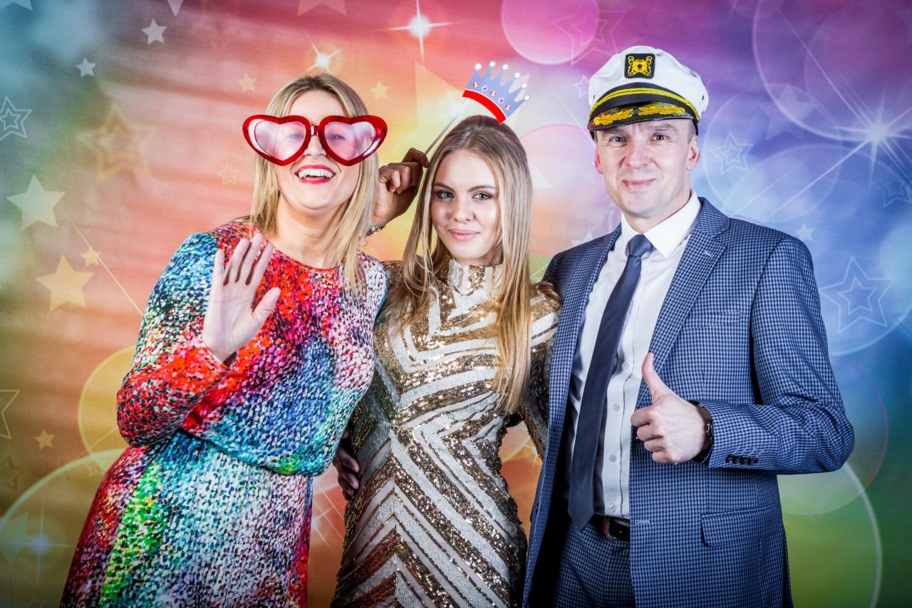 Trzy osoby pozują na tle kolorowego tła z gwiazdami: kobieta w tęczowej sukience i okularach w kształcie serc macha ręką, kobieta w złotej sukience trzyma papierową koronę, mężczyzna w marynarskim...