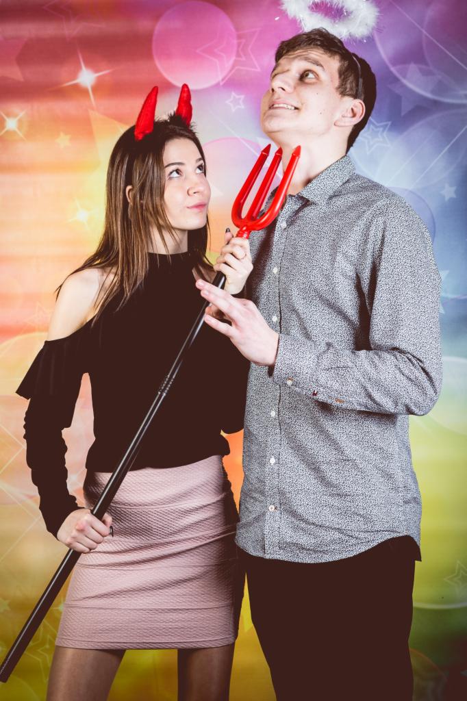 Para w przebraniach: kobieta z rogami diabła trzyma trójząb przy szyi mężczyzny z aureolą na głowie, na tle kolorowego tła z gwiazdami i okręgami.