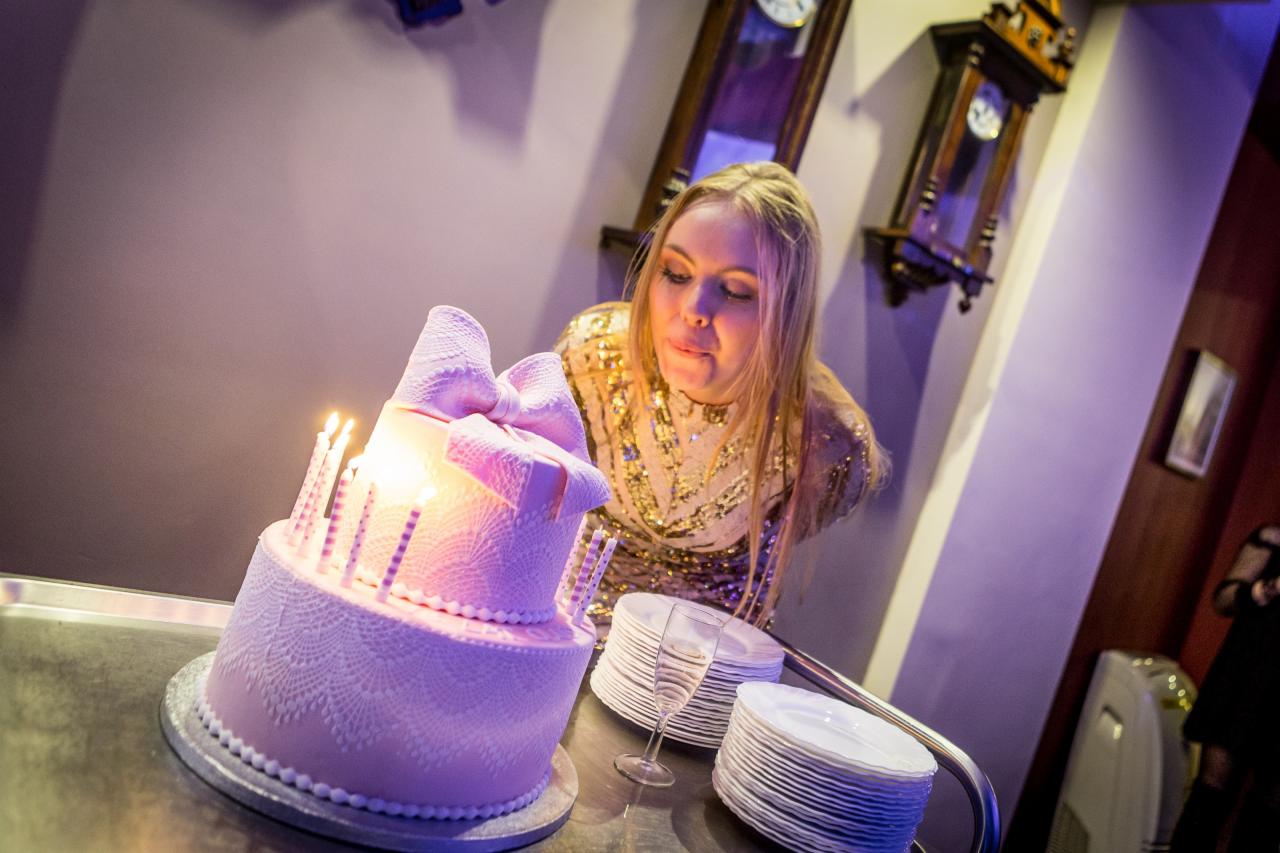 Kobieta w złotej sukience dmucha świeczki na dwupiętrowym, różowym torcie ozdobionym koronką i kokardą, obok stosy białych talerzy i kieliszek do szampana, w tle wiszące zegary.