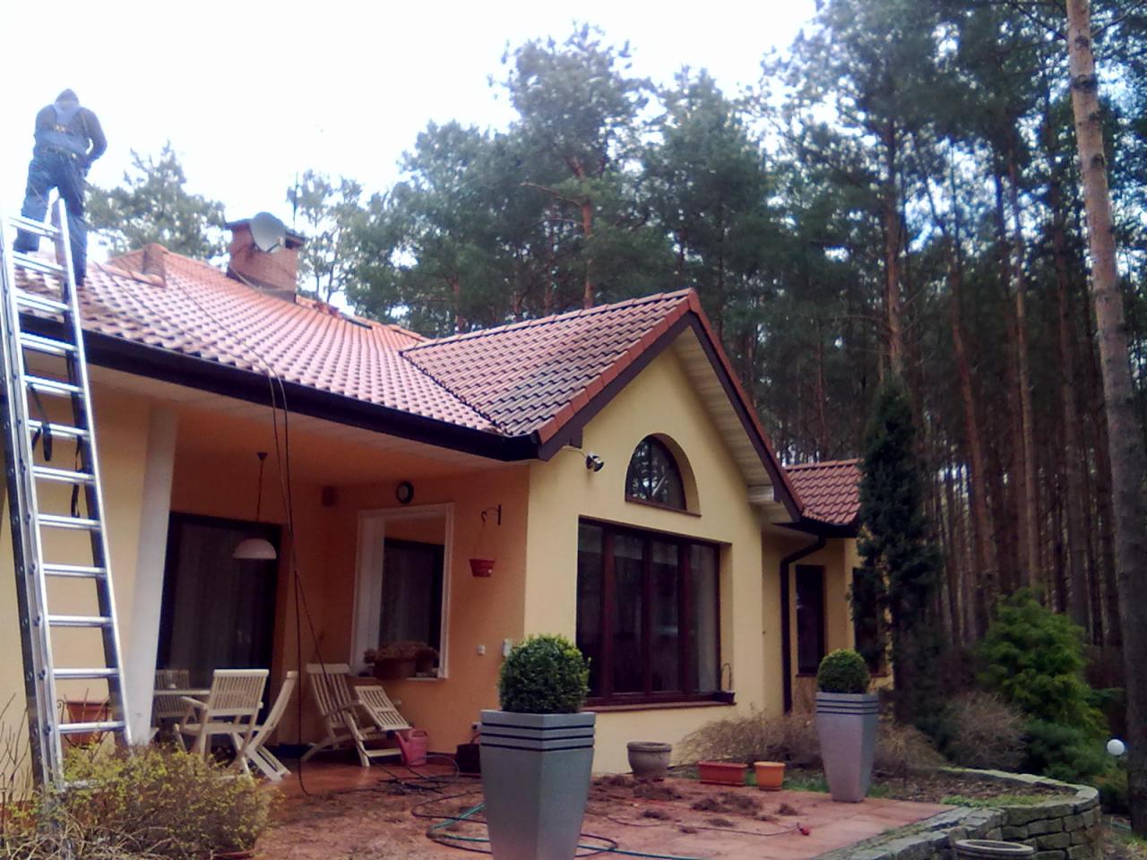 Pracownik na drabinie przy domu z pomarańczową elewacją i czerwoną dachówką, prawdopodobnie wykonujący prace na wysokości, z widocznym lasem w tle.
