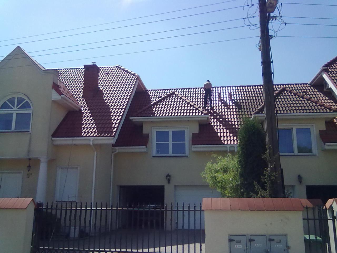 Pracownik w ciemnych spodniach i jasnej koszulce na dachu domu z czerwoną dachówką, widok z dołu na tle błękitnego nieba i słupa energetycznego.