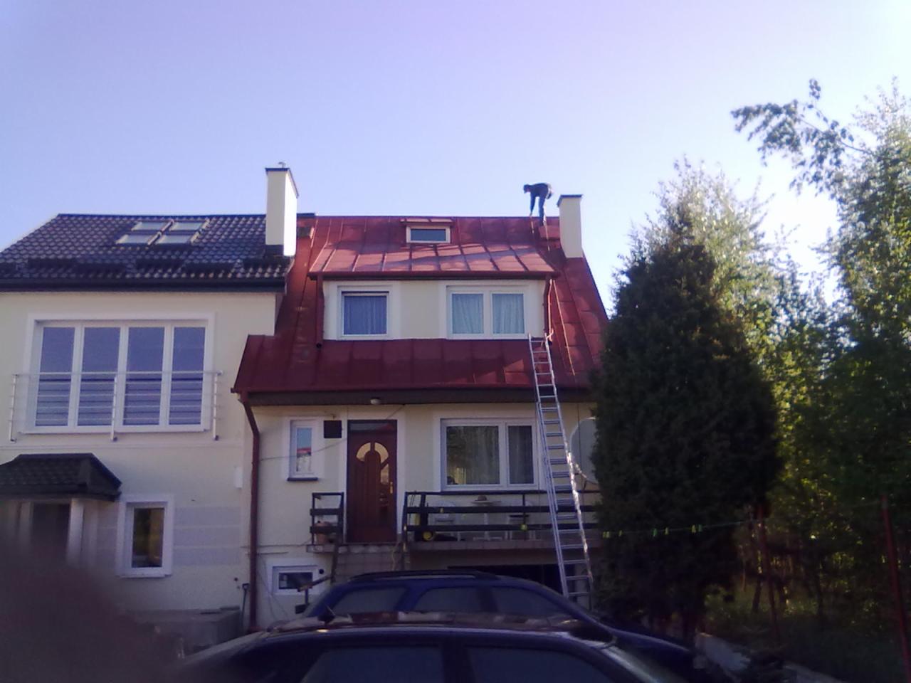 Prace wysokościowe na dachu dwupiętrowego domu z czerwoną blachodachówką, widoczna drabina oparta o elewację i sylwetka pracownika na szczycie komina.