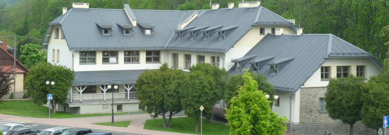 Wielopiętrowy budynek z szarą blachodachówką, widok z góry na elewację i otoczenie z drzewami i parkingiem.