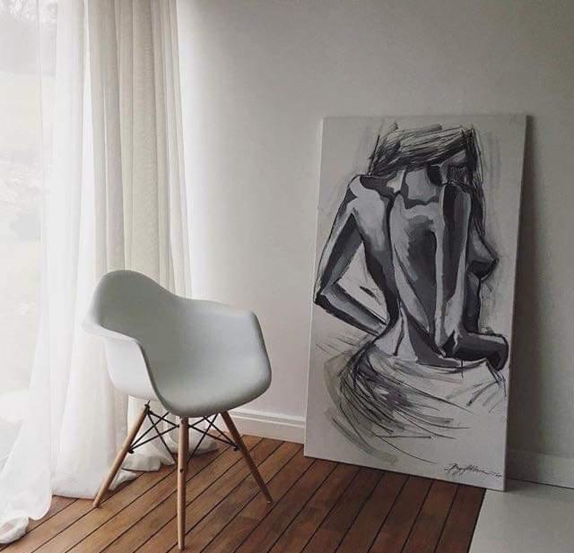 Szkic nagiej kobiety na płótnie oparty o białą ścianę, obok białe krzesło Eames na drewnianej podłodze, w tle zasłonięte okno.