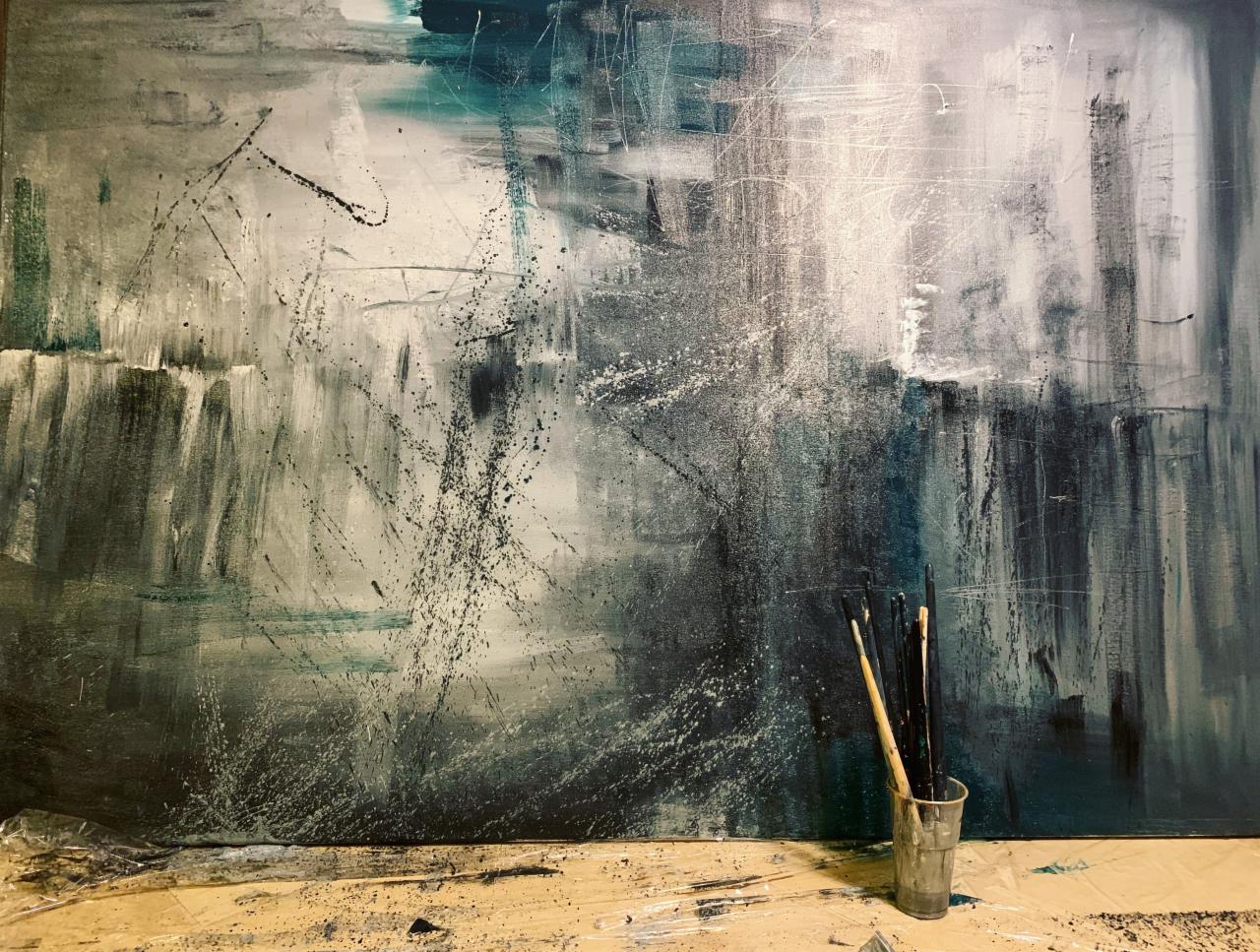 Abstrakcyjne malowidło w odcieniach bieli, szarości i granatu z widocznymi pociągnięciami pędzla i drobnymi odpryskami farby, w prawym dolnym rogu plastikowy kubek z kilkoma pędzlami.