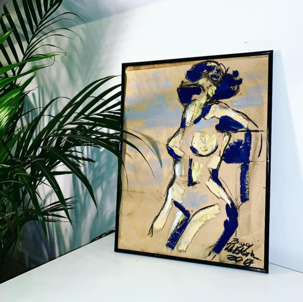 Szkic aktu kobiecego w ramie, wykonany farbami w odcieniach błękitu, bieli i złota, ustawiony na białym blacie obok zielonej rośliny doniczkowej.