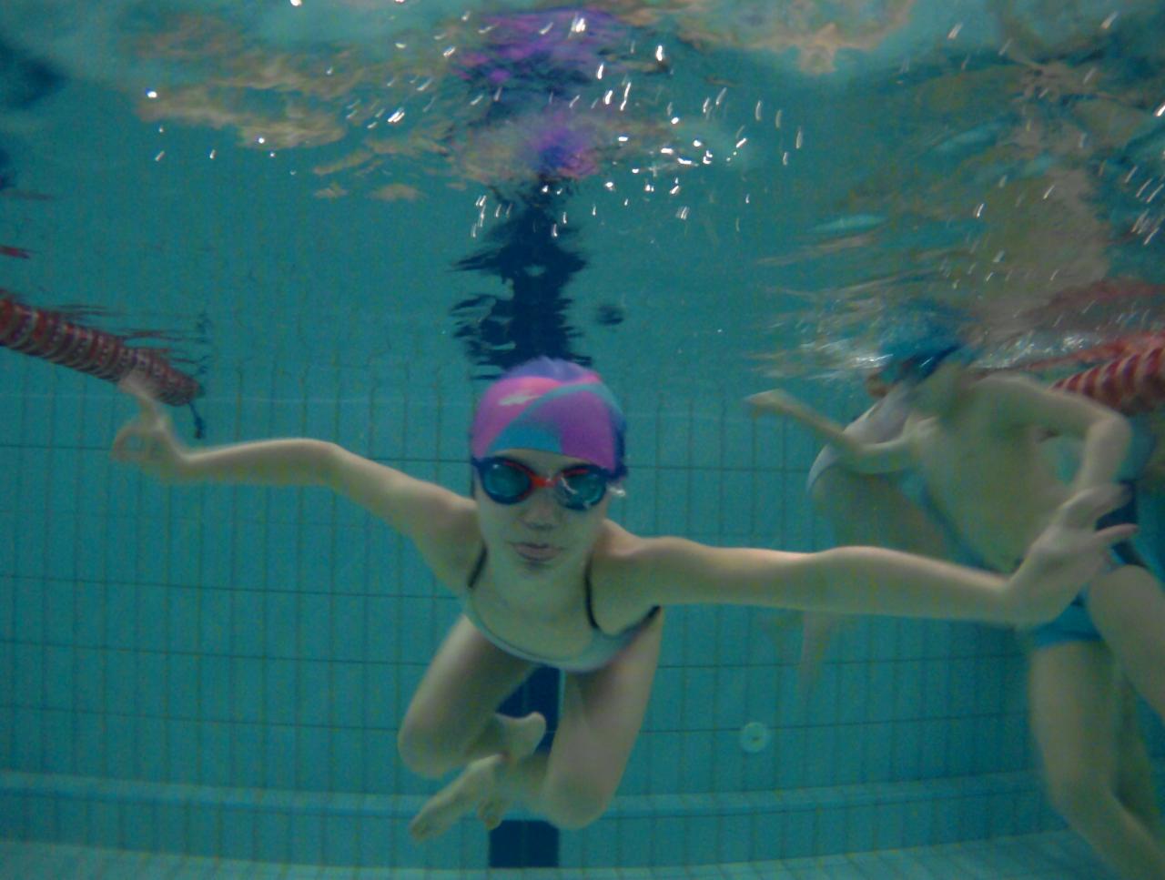 Dziewczynka w różowo-niebieskim czepku i okularach pływackich unosi się pod wodą w basenie, z rozłożonymi ramionami, widok z dołu, lekko zamazane tło z innymi pływającymi dziećmi.