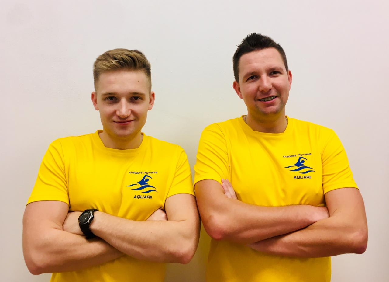 Dwóch instruktorów w żółtych koszulkach z logo Akademii Pływania Aquarii, jeden z zegarkiem na ręce, drugi z aparatem ortodontycznym, pozują z założonymi rękami na tle jasnej ściany.