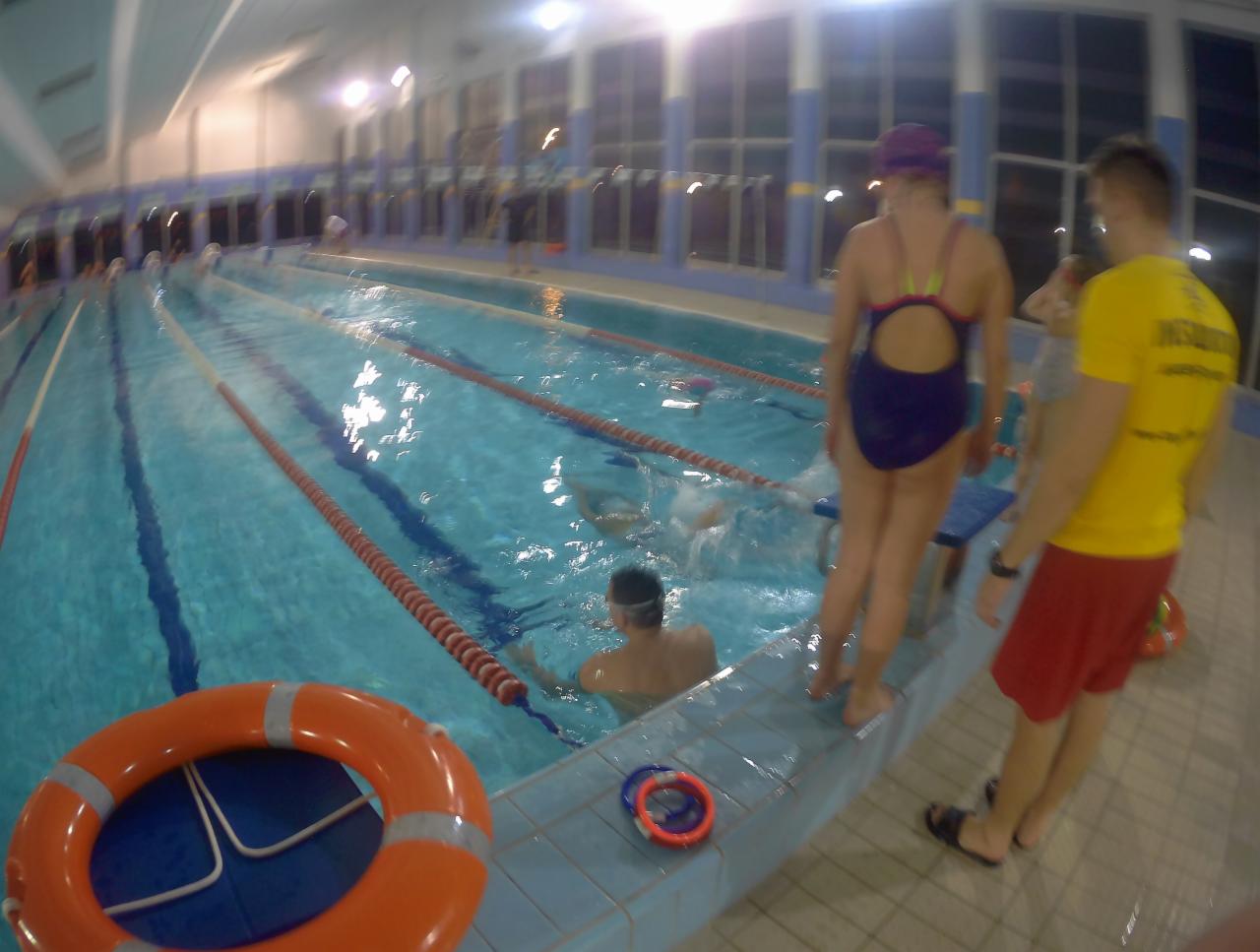 Kryty basen z osobami pływającymi i instruktorem w żółtej koszulce oraz pomarańczowe koło ratunkowe na pierwszym planie.