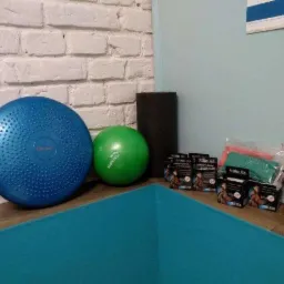 Niebieska poduszka sensoryczna, zielona piłka, wałek piankowy, opakowania taśm kinezjologicznych i spray Pre-Tape na tle ściany z białej cegły i turkusowej maty do ćwiczeń.