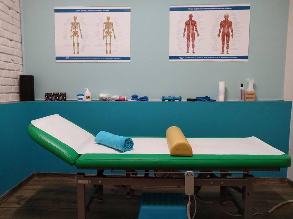 Zielony stół do masażu z niebieskim ręcznikiem i żółtym wałkiem, plakaty z układem kostnym i mięśniowym na ścianie, półka z akcesoriami do masażu w tle.