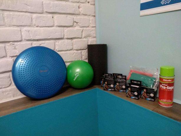 Niebieska poduszka sensoryczna, zielona piłka, wałek piankowy, opakowania taśm kinezjologicznych i spray Pre-Tape na tle ściany z białej cegły i turkusowej maty do ćwiczeń.