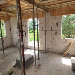 Wnętrze surowego domu w trakcie budowy, ściany z białego betonu komórkowego, drewniane szalunki stropu podparte metalowymi stemplami, widok na zieleń za oknami.