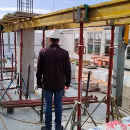 Pracownik w kasku obserwuje postęp prac na budowie, widoczne szalunki stropowe, podpory, bloczki betonowe i dźwig nożycowy w tle.