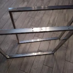 Dwie stalowe, spawane ramy stołu z nawierconymi otworami na blacie, leżące na podłodze z paneli imitujących drewno.