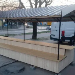 Zadaszona konstrukcja handlowa z drewna i metalu, dwupoziomowa, umieszczona na chodniku, w tle biały bus z otwartymi drzwiami.