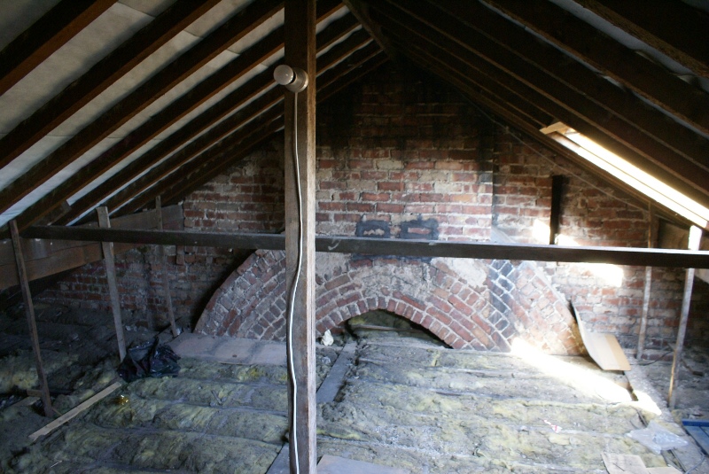Nieocieplone poddasze z widocznymi krokwiami, starą ceglaną ścianą kominową z łukowym otworem i częściowo rozłożoną izolacją z wełny mineralnej.