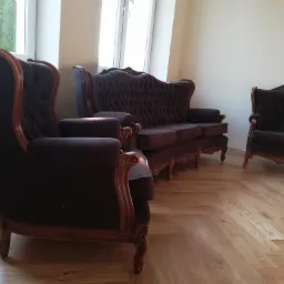 Komplet wypoczynkowy w stylu Ludwika Filipa: sofa trzyosobowa i dwa fotele uszaki z ciemną tapicerką i rzeźbionymi drewnianymi ramami, ustawione na parkiecie w jodełkę przy jasnych ścianach i oknie.