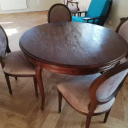 Okrągły, ciemnobrązowy stół z krzesłami o jasnej, pluszowej tapicerce i drewnianych ramach. Parkiet w jodełkę na podłodze.