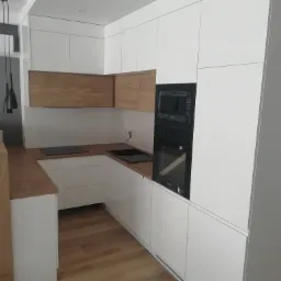 Nowoczesna kuchnia z białymi, lakierowanymi frontami i drewnianym blatem, czarne AGD w zabudowie, minimalistyczne lampy wiszące.