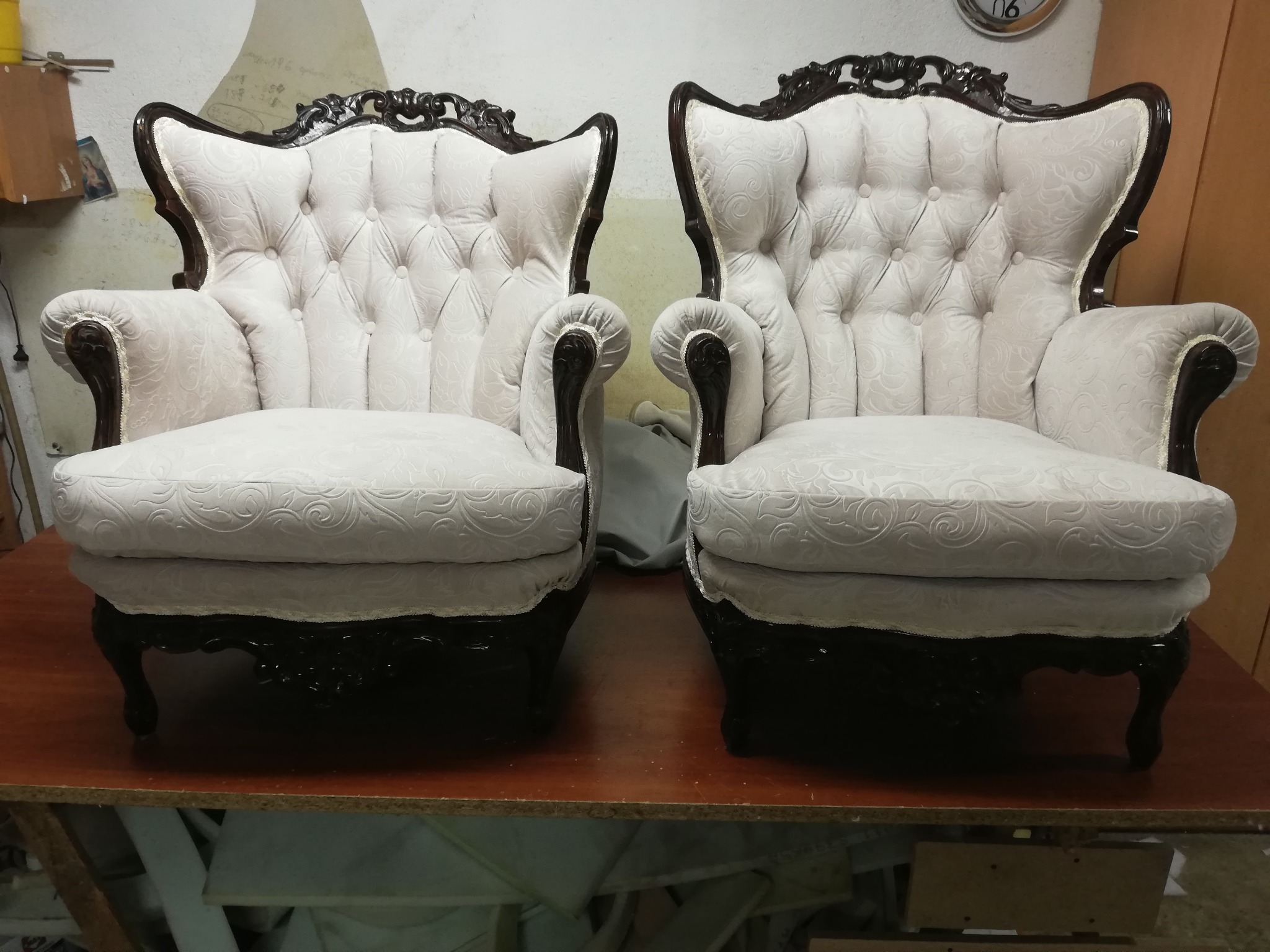 Dwa identyczne, białe fotele klubowe z ciemnobrązowymi, rzeźbionymi elementami drewnianymi, ustawione obok siebie na stole w warsztacie, widoczne ślady pracy w tle.