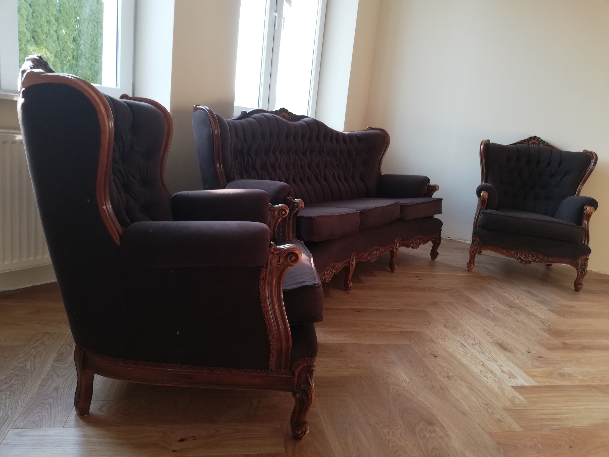 Komplet wypoczynkowy w stylu Ludwika Filipa: sofa trzyosobowa i dwa fotele uszaki z ciemną tapicerką i rzeźbionymi drewnianymi ramami, ustawione na parkiecie w jodełkę przy jasnych ścianach i oknie.