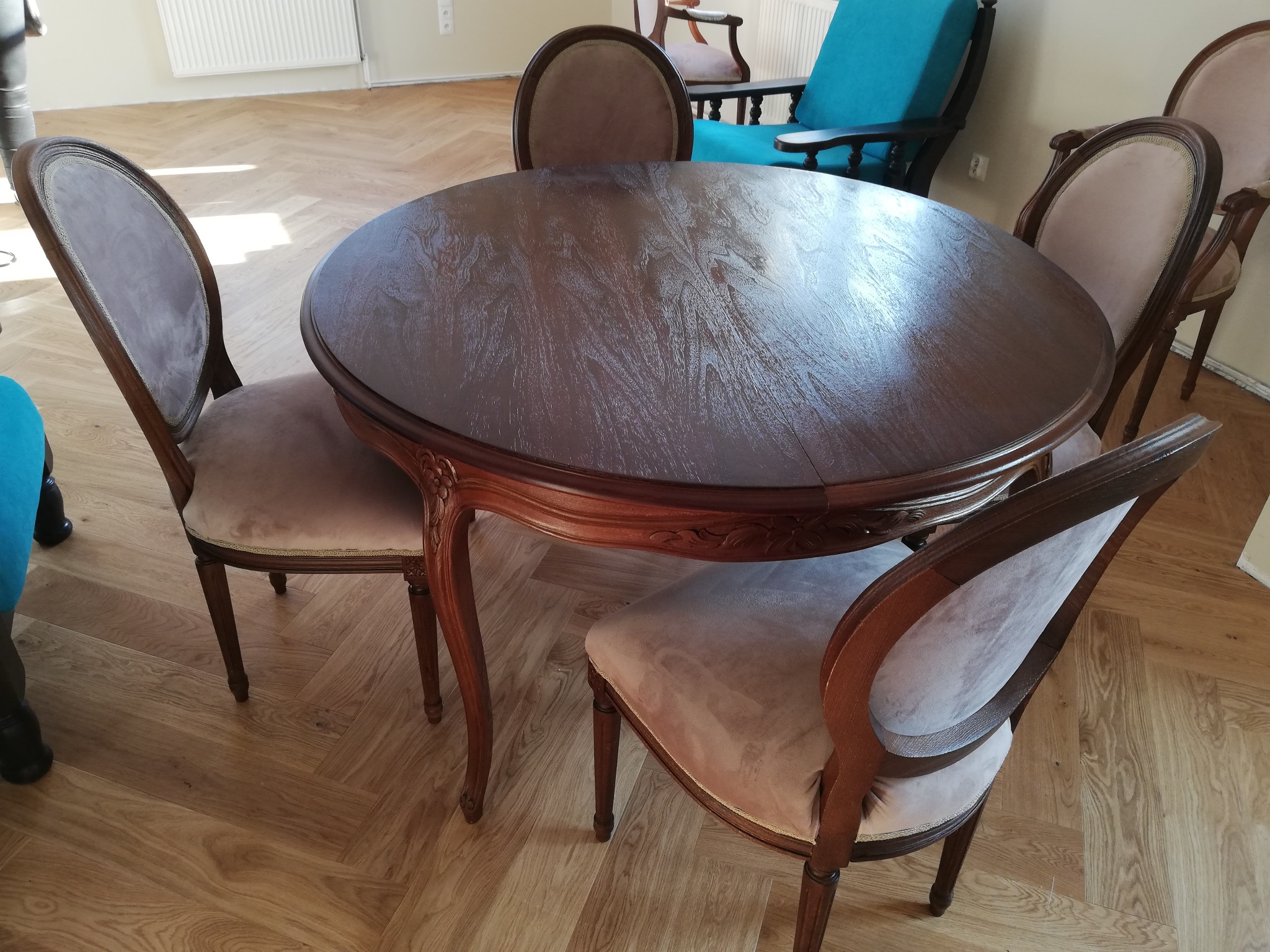 Okrągły, ciemnobrązowy stół z krzesłami o jasnej, pluszowej tapicerce i drewnianych ramach. Parkiet w jodełkę na podłodze.