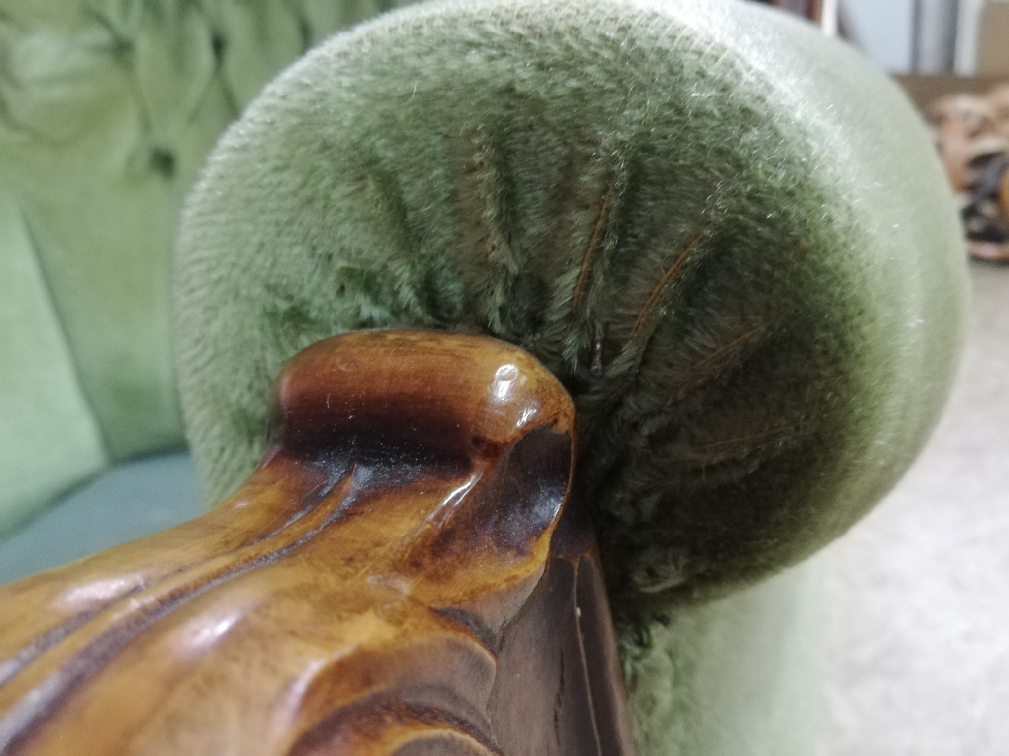 Detail of a light green, plush upholstered armchair armrest with visible stitching and a dark wood carved arm, showing signs of wear.