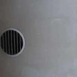 Okrągła kratka wentylacyjna z metalowymi prętami zamontowana na szarej, lekko zabrudzonej ścianie, widoczna obok czarnej, żaluzjowej powierzchni.