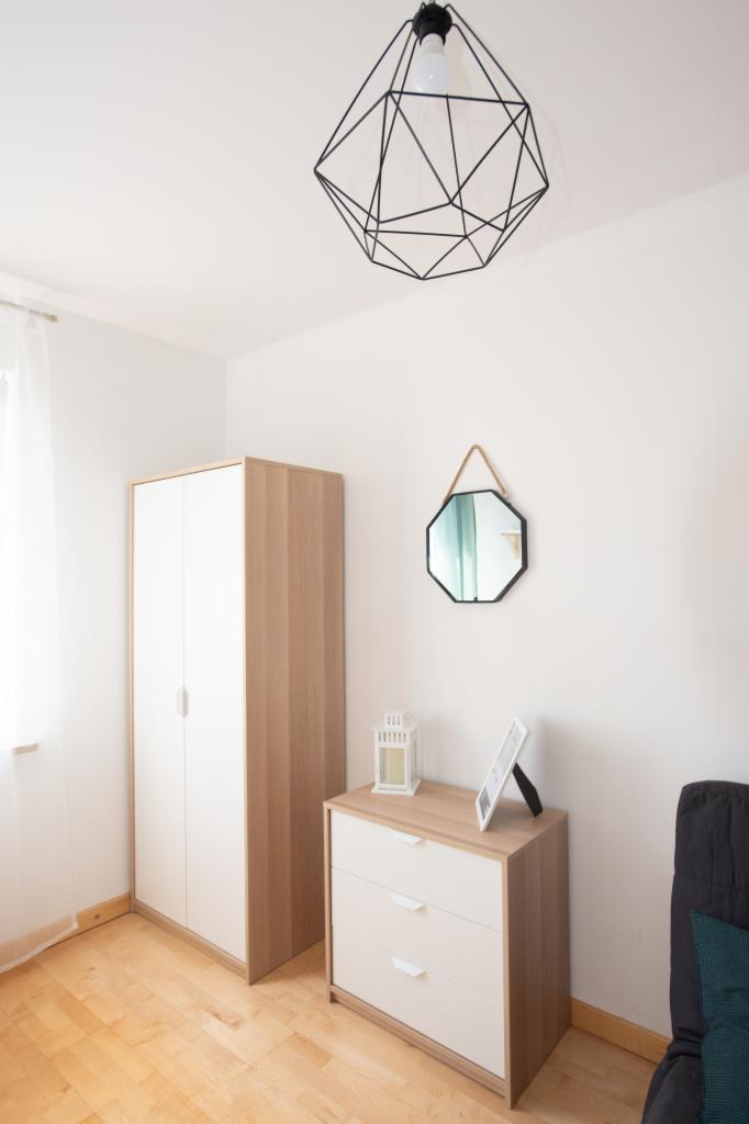 Minimalistyczna sypialnia z drewnianą podłogą, białą szafą, komodą z trzema szufladami, ośmiokątnym lustrem na sznurku i geometryczną lampą wiszącą.