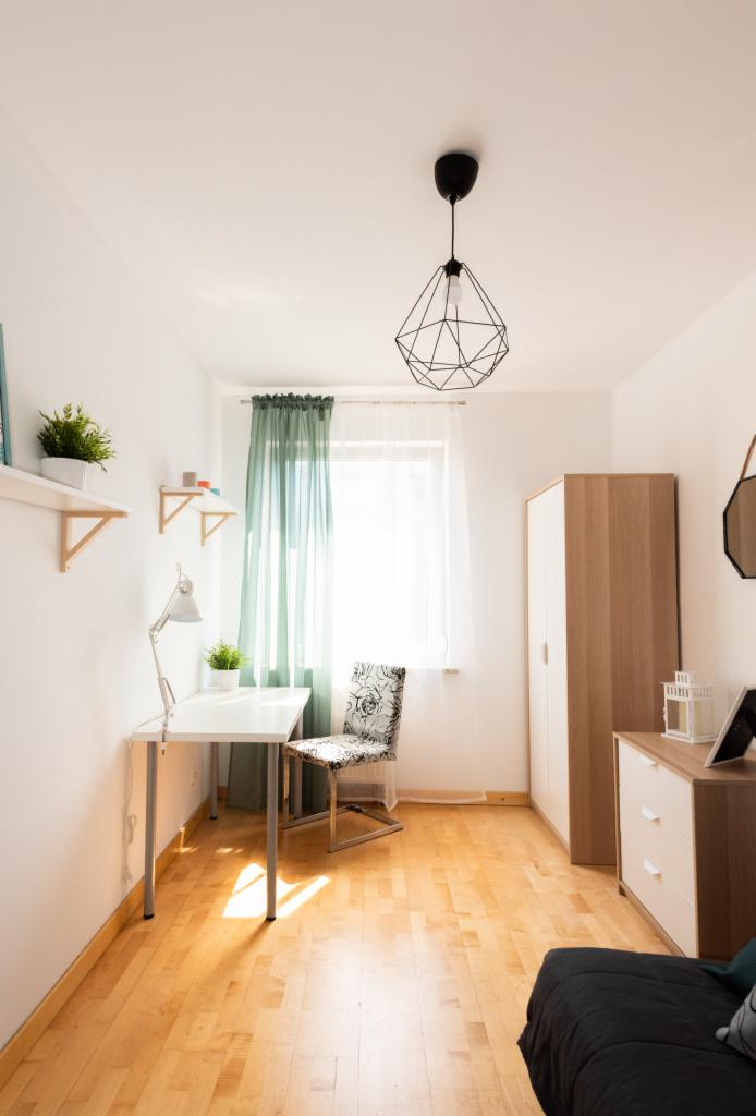 Jasne wnętrze pokoju z biurkiem, krzesłem w kwiaty, szafą i lampą geometryczną; drewniana podłoga i zasłony w odcieniach zieleni.