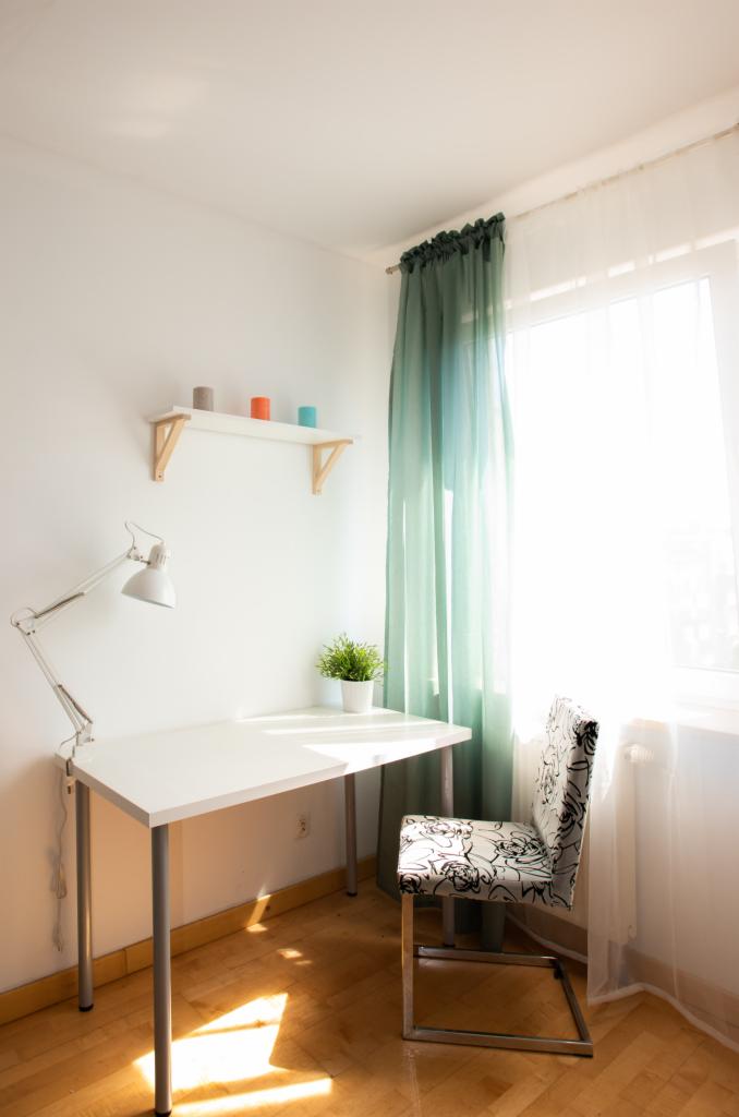 Jasny pokój z biurkiem, lampką, krzesłem w kwiaty i turkusowymi zasłonami, oświetlony promieniami słońca wpadającymi przez okno.