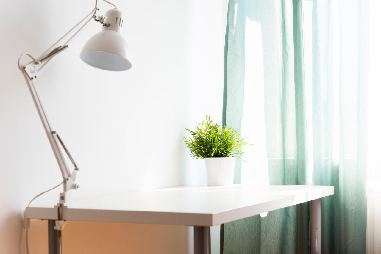Białe biurko z regulowaną lampą i doniczką z zieloną rośliną na tle okna z turkusową zasłoną.