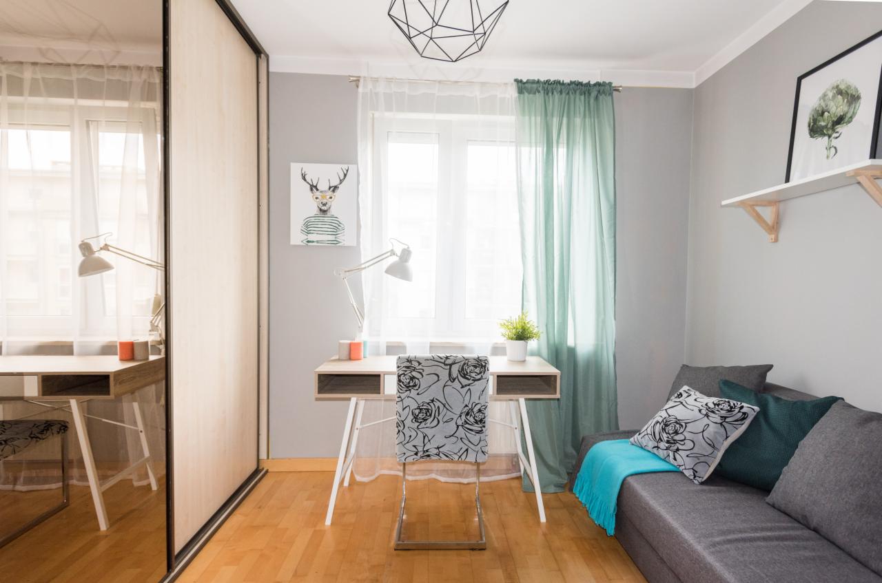 Jasne wnętrze pokoju z biurkiem, krzesłem w kwiaty, szarą sofą z turkusowym pledem i geometryczną lampą sufitową.