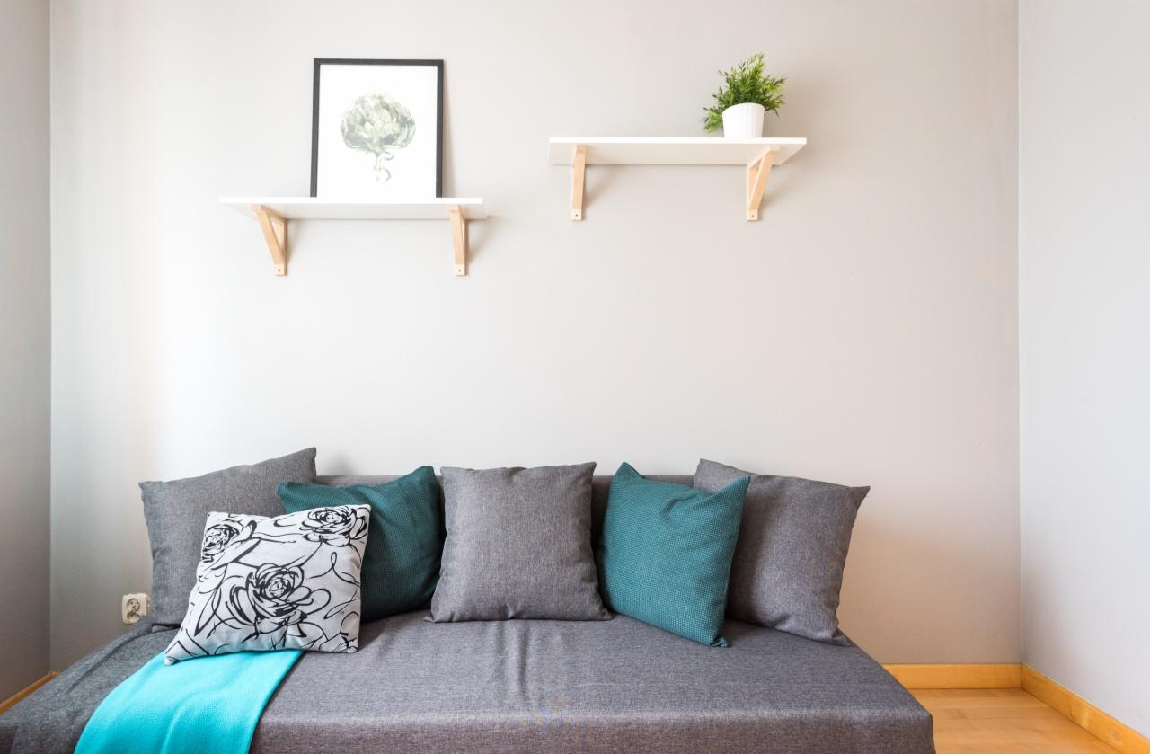 Szary tapczan z poduszkami w odcieniach szarości i turkusu, przykryty turkusowym pledem. Na ścianie dwie białe półki z drewnianymi wspornikami: na jednej roślina w doniczce, na drugiej grafika...