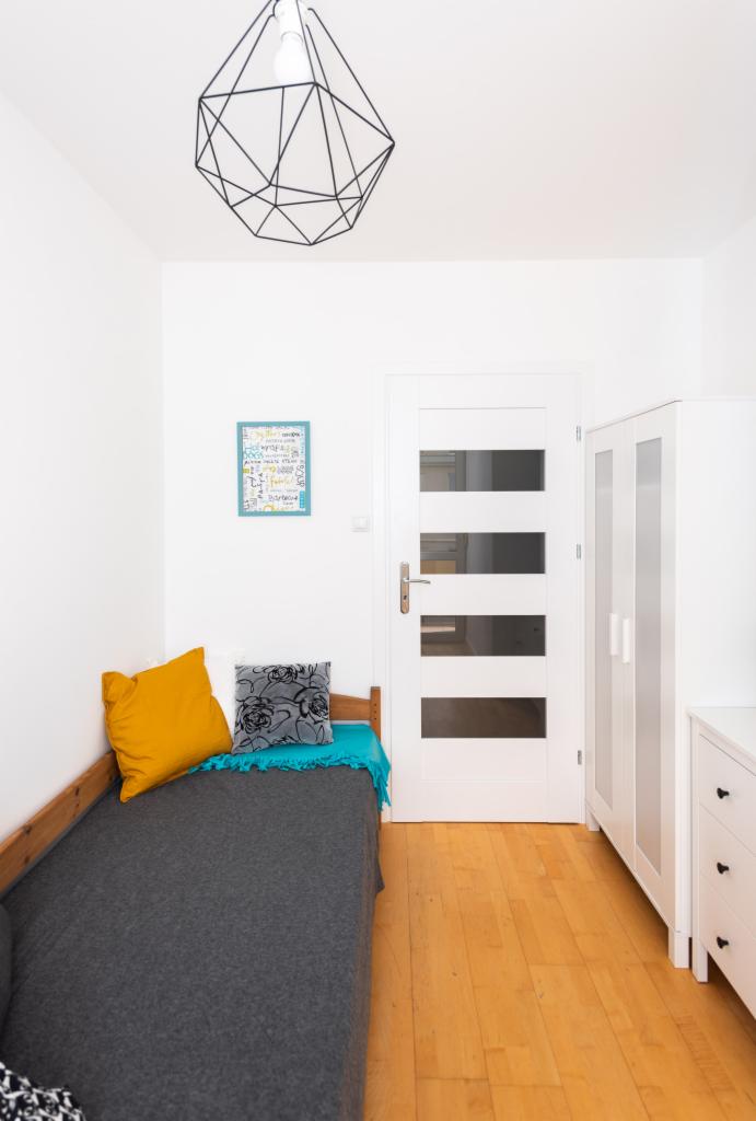 Pokój z pojedynczym łóżkiem, żółtą poduszką i geometryczną lampą wiszącą, białe ściany, drzwi z przeszkleniami i drewniana podłoga.