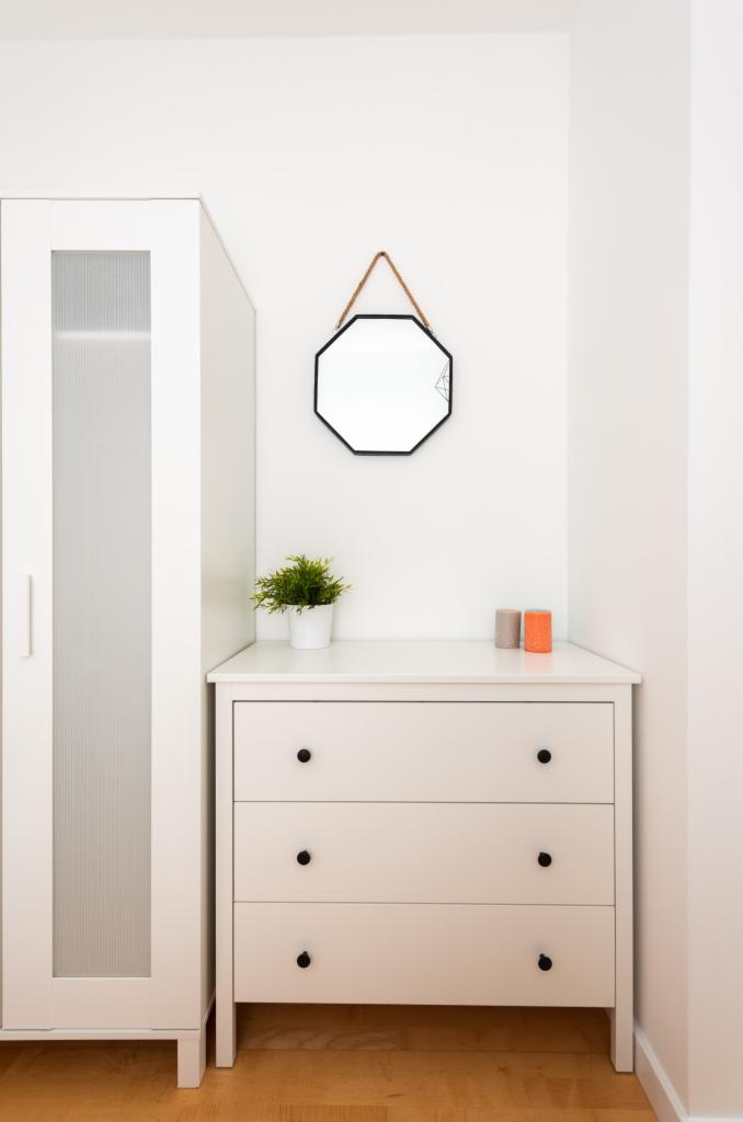 Biała komoda z trzema szufladami i czarnymi gałkami, obok biała szafa z przeszklonym frontem, nad komodą ośmiokątne lustro w czarnej ramie zawieszone na sznurku, mała roślina doniczkowa i dwie...