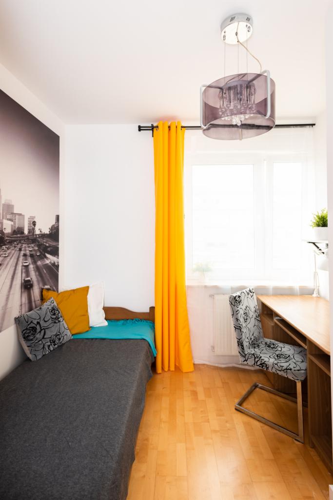 Pokój młodzieżowy z pojedynczym łóżkiem, biurkiem i fototapetą z motywem miejskim, oświetlony lampą z fioletowymi akcentami i pomarańczowymi zasłonami.