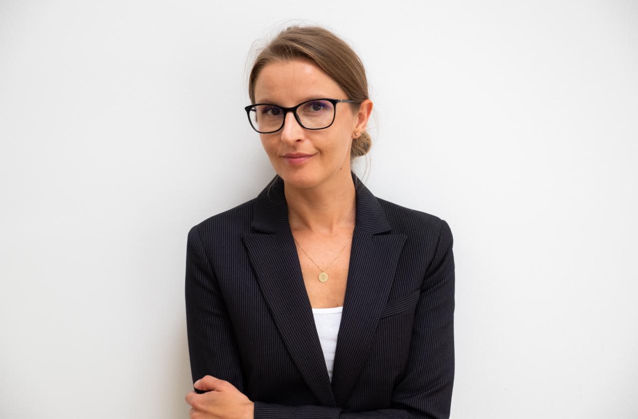 Kobieta w okularach i ciemnym garniturze na tle białej ściany, z delikatnym uśmiechem i złotym naszyjnikiem.