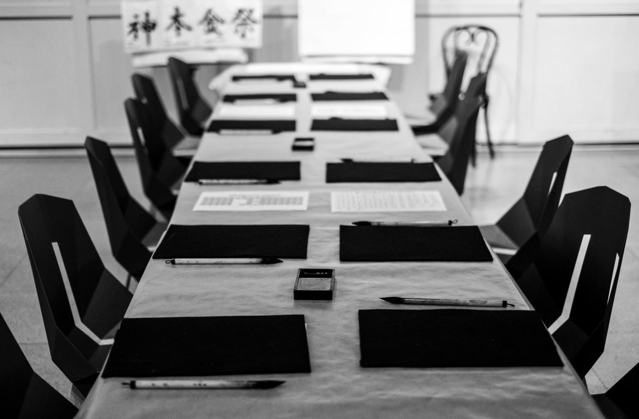 Długi stół nakryty jasnym obrusem, przygotowany do kaligrafii: czarne podkładki, pędzle, kałamarze z tuszem i kartki z tekstem. Czarno-biała fotografia.