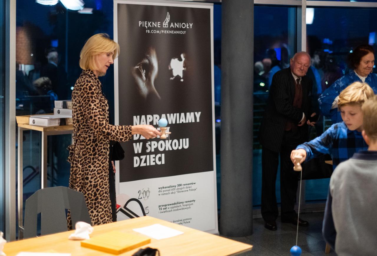 Kobieta w sukience w panterkę trzyma kendamę przed plakatem fundacji 'Piękne Anioły', w tle widoczni ludzie i bawiące się dzieci.