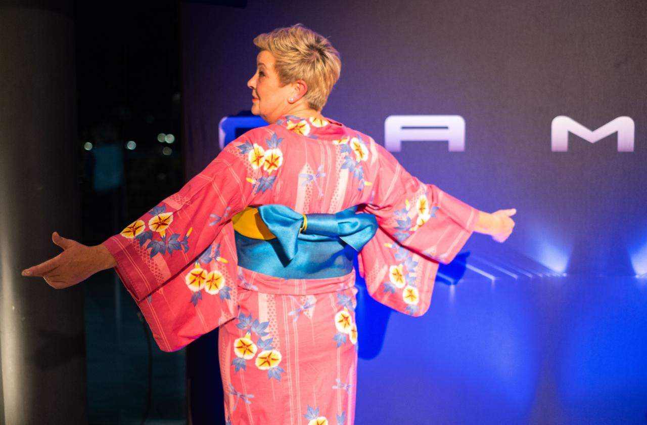 Kobieta w różowym kimonie z niebieskim pasem, stojąca tyłem na tle ciemnej ściany z jasnymi literami.