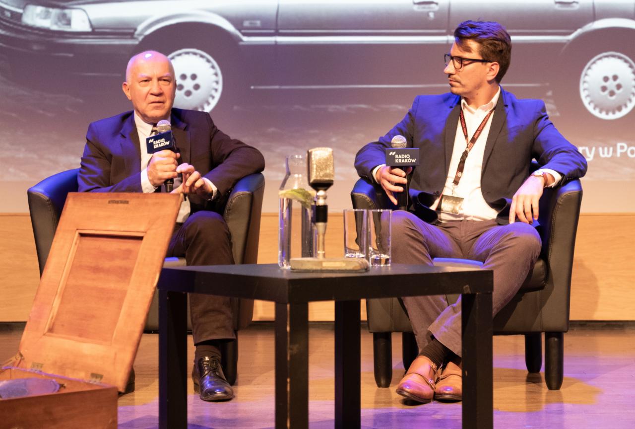 Dwóch mężczyzn w garniturach siedzi na krzesłach, prowadząc wywiad z mikrofonami Radio Kraków, na tle ekranu z samochodem i otwartą drewnianą skrzynką obok jednego z nich.
