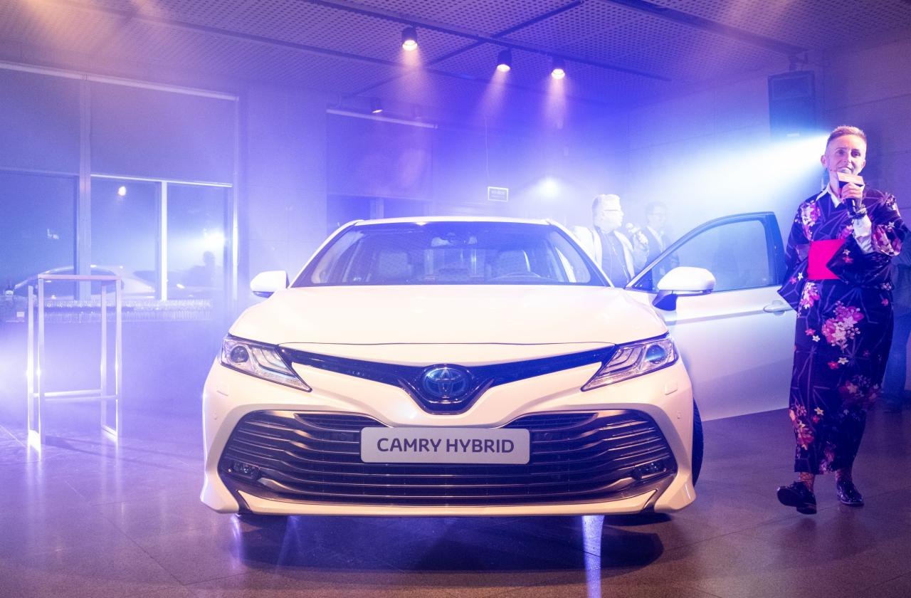 Biały samochód Camry Hybrid na prezentacji, kobieta w kwiecistym kimonie z mikrofonem, niebieskie oświetlenie sceniczne, goście w tle.