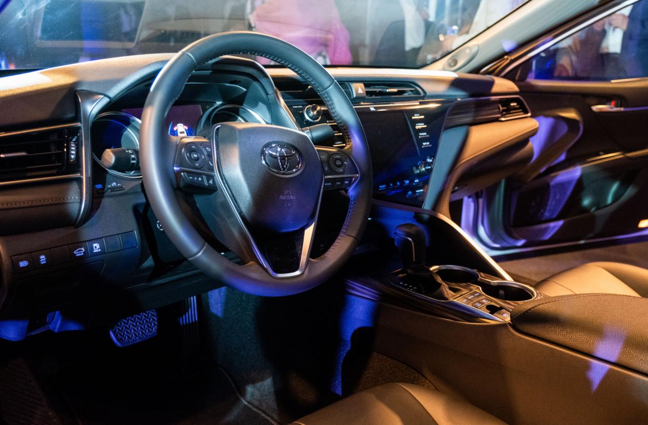Wnętrze samochodu Toyota Camry z widoczną deską rozdzielczą, kierownicą z logo i elementami podświetlenia w kolorze niebieskim.